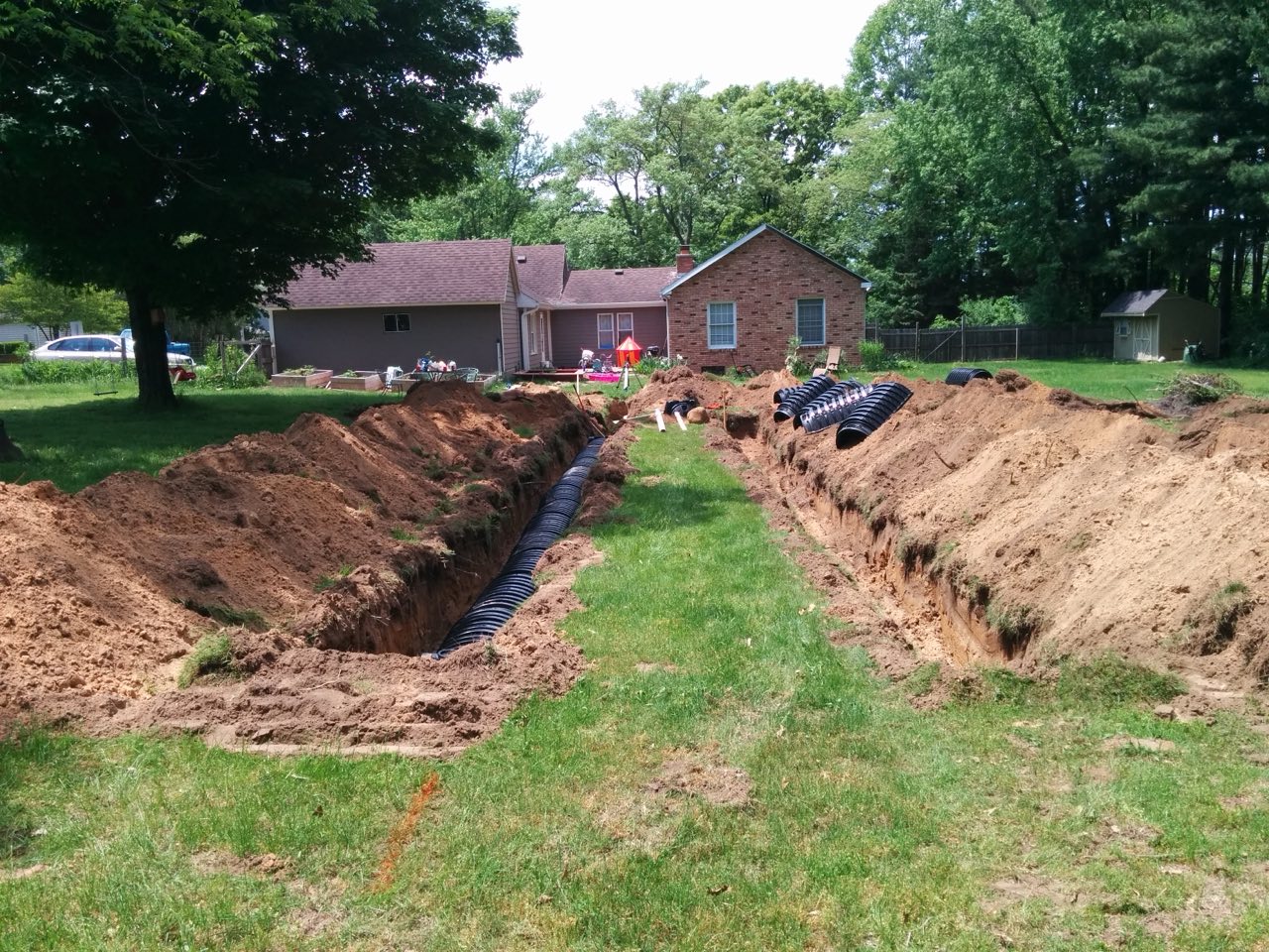 Two freshly dug drain field trenches, one holding black plastic chambers, across a residential backyard with a house behind.