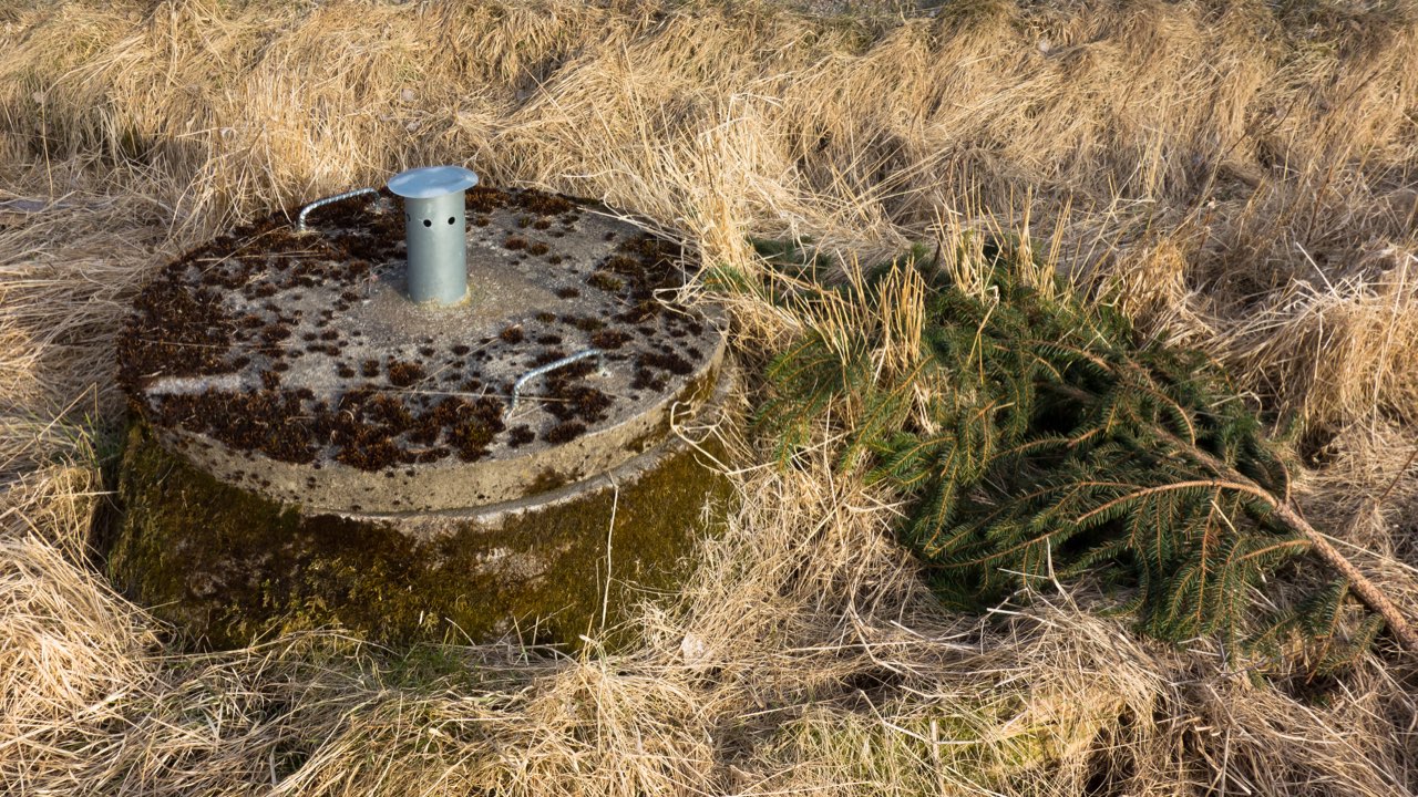 A round concrete septic tank lid with a short metal vent pipe, set in dry grass.